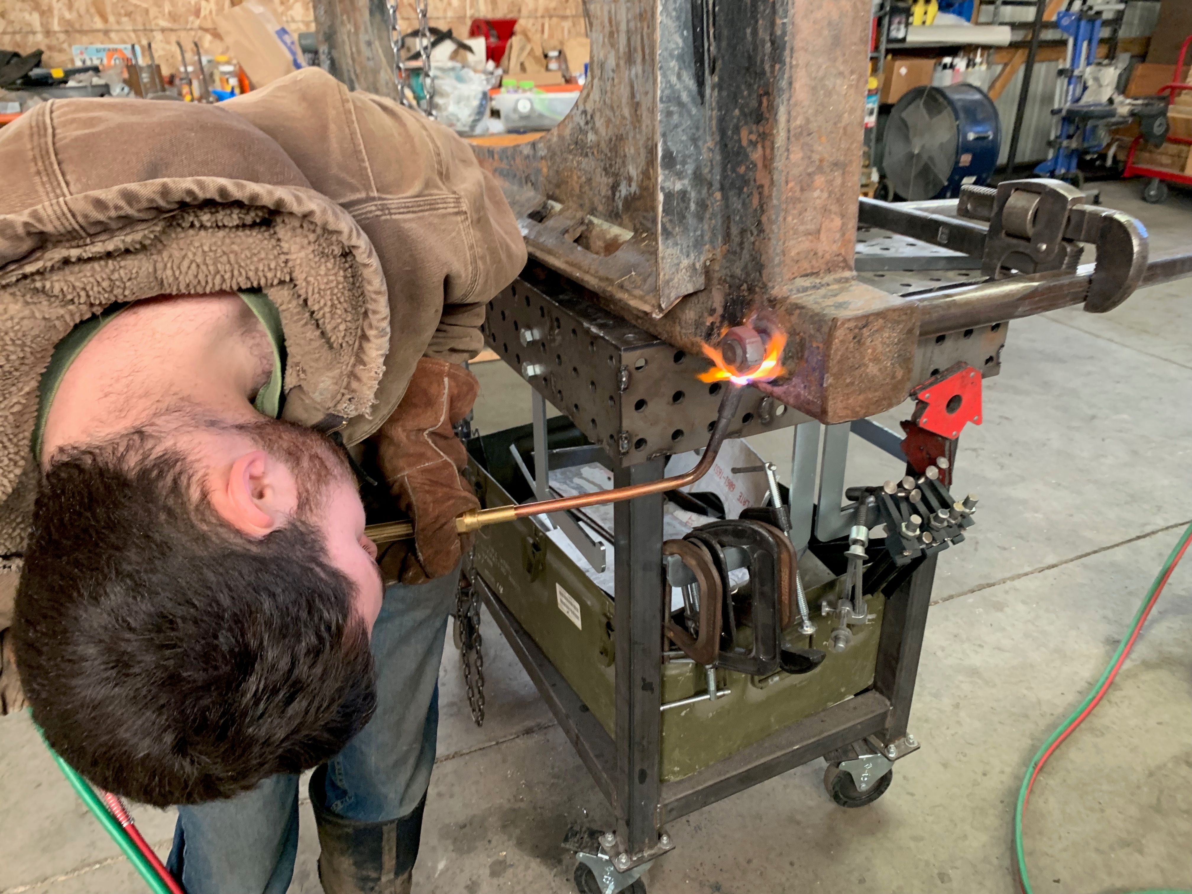 Jimmie with his head upside-down, using a torch to loosen a rusted bolt on a large hay fork for a tractor.